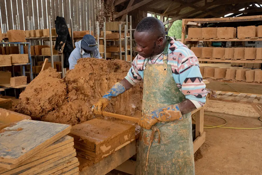 Dans un atelier sous charpente, un artisan en tablier tasse de la terre dans un moule à briques ; des briques sèchent sur des étagères