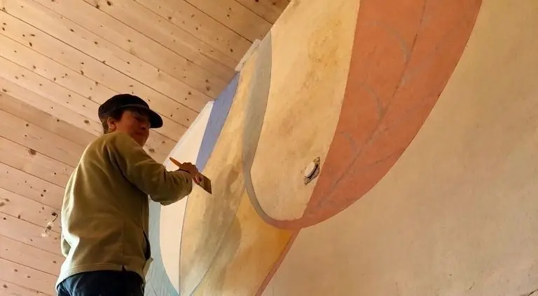 Une artisane peint un grand décor arrondi sur un mur enduit, sous un plafond en lambris