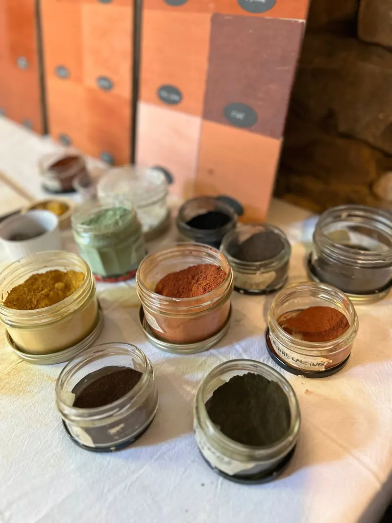 Bocaux de pigments en poudre, ocre, rouge, vert et noir, devant un nuancier d'enduits